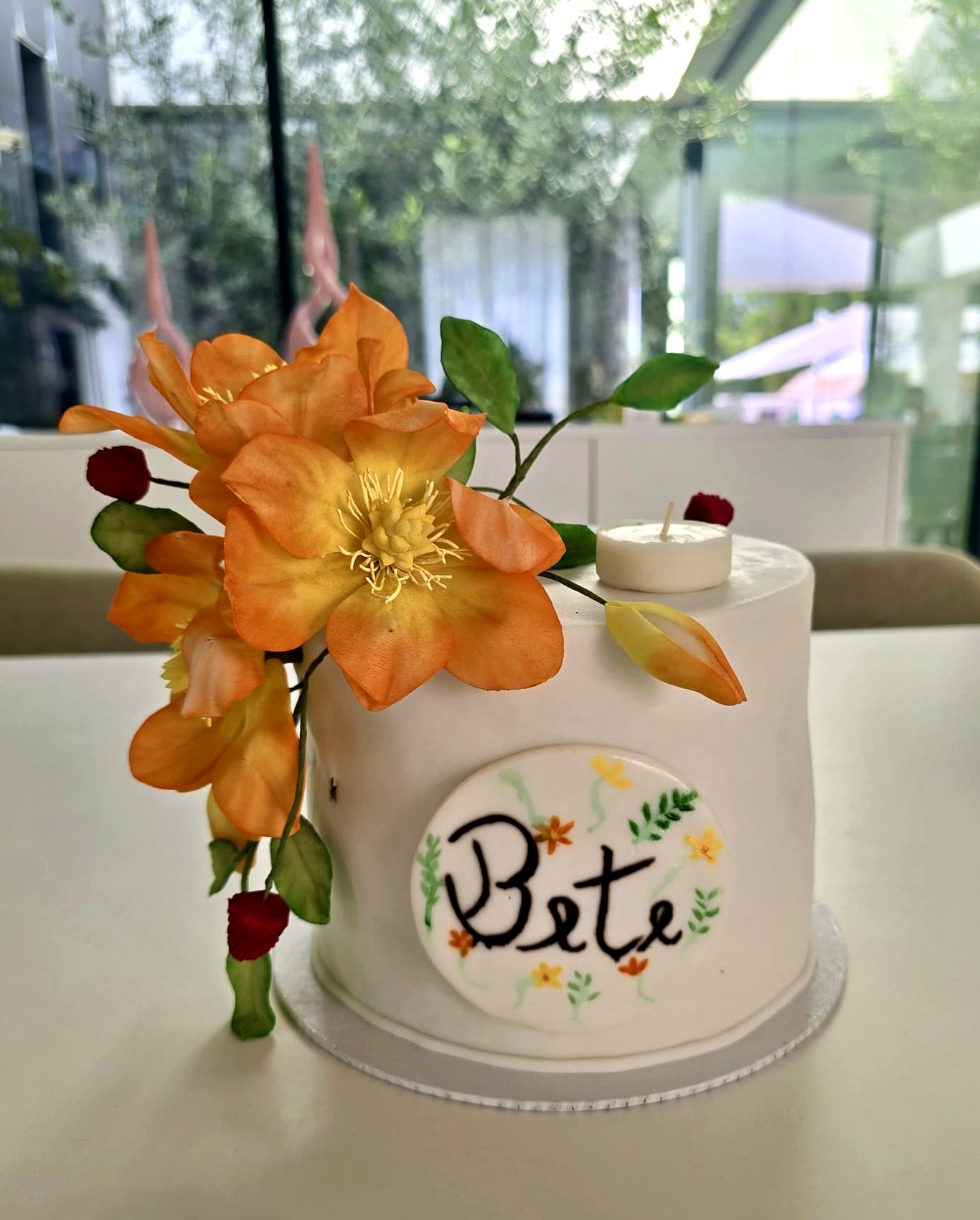 Tarta blanca con flor naranja grande, detalles rojos y placa con el nombre Bete.