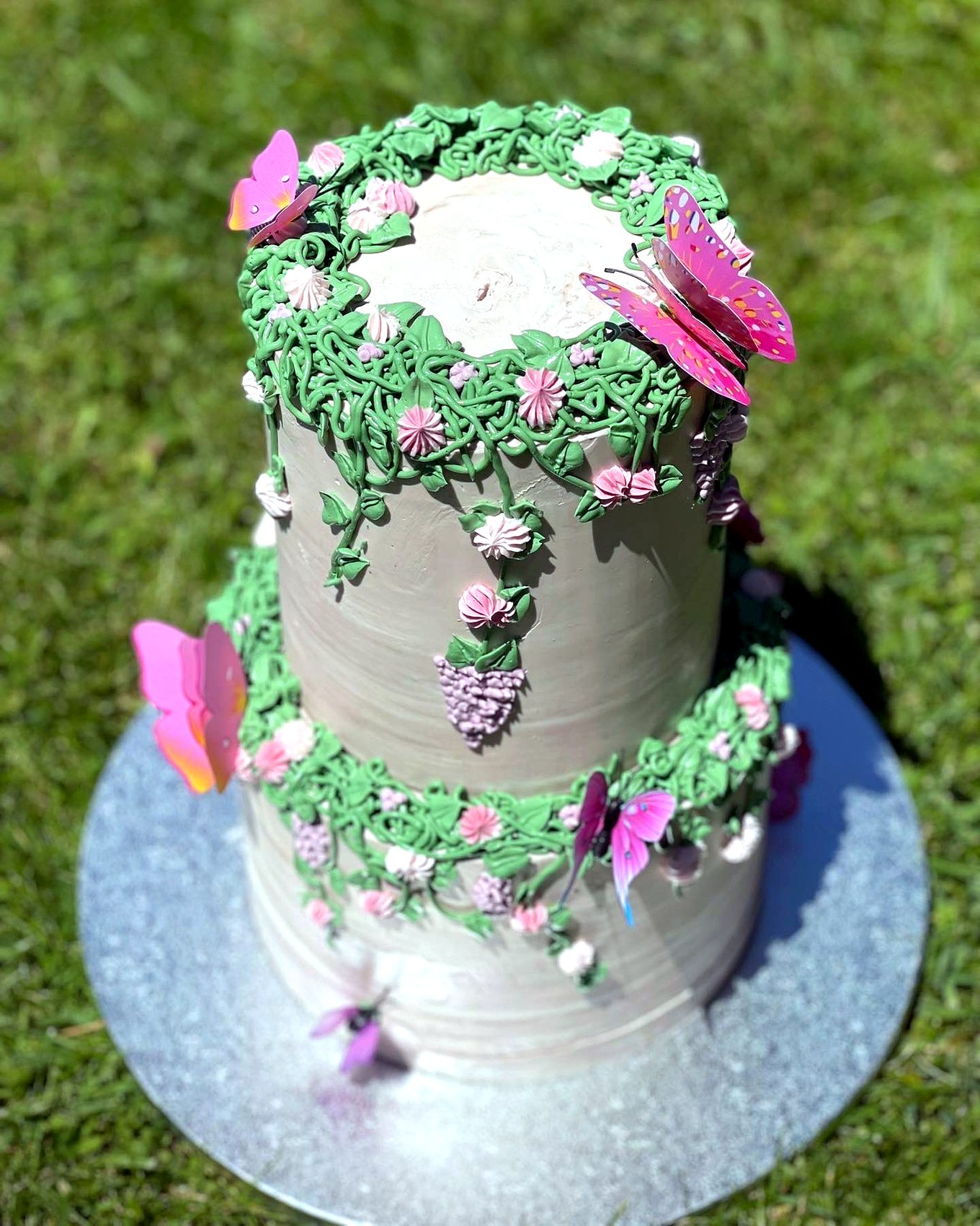 Tarta de dos niveles con enredaderas, flores rosas y mariposas sobre cesped.
