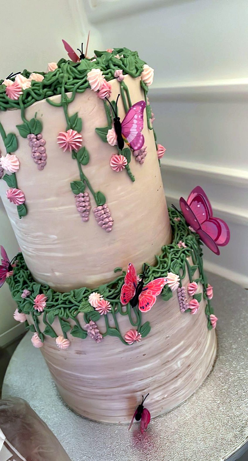 Tarta inclinada de dos niveles con mariposas rosas, flores y enredaderas verdes.