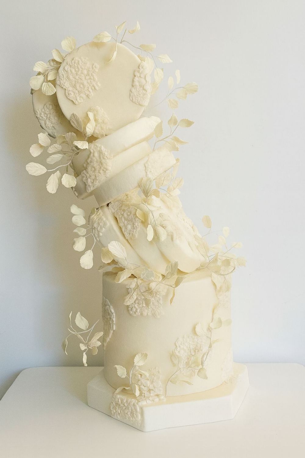 Tarta alta blanca con arreglo diagonal de flores y hojas claras sobre base cuadrada.
