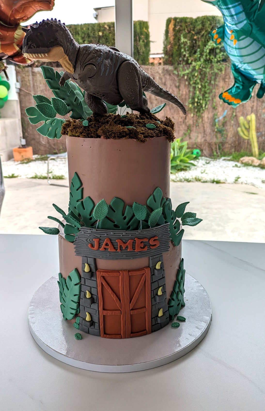 Detalle de tarta de dinosaurios con figura superior, hojas tropicales y nombre James.