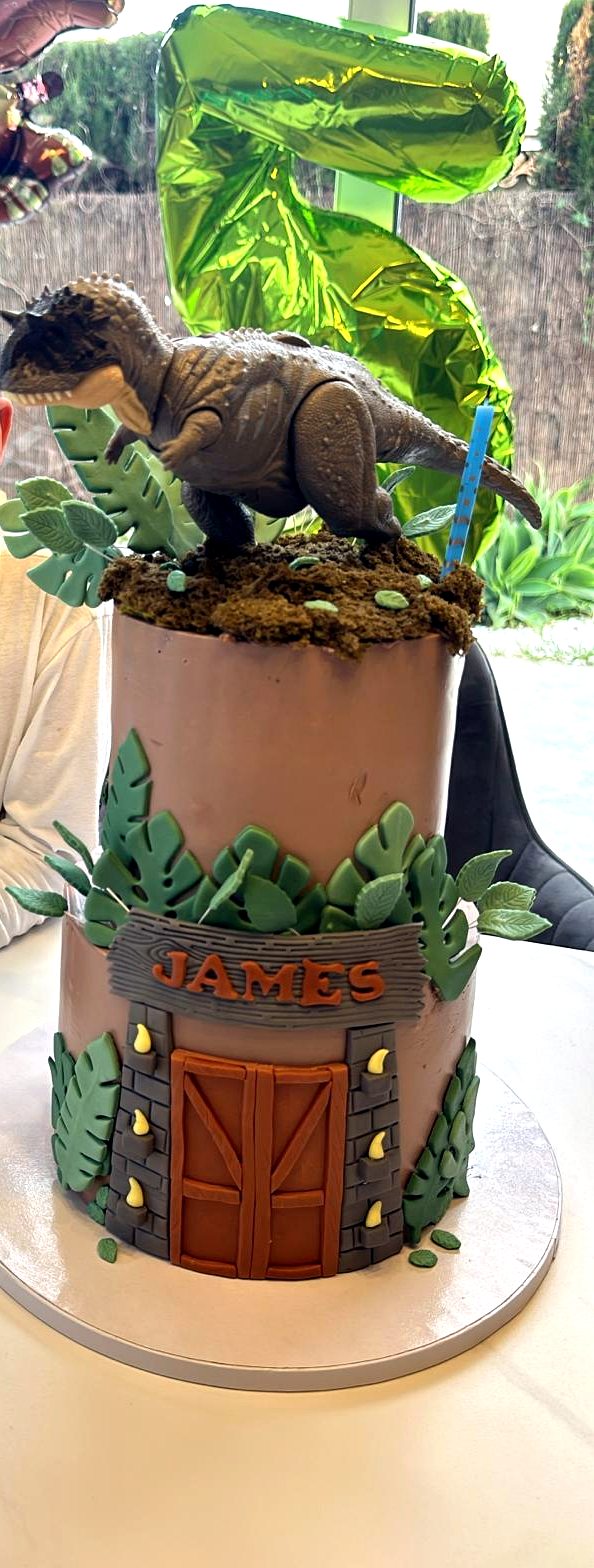 Tarta de dinosaurios con nombre James, puerta de parque, hojas y figura superior.