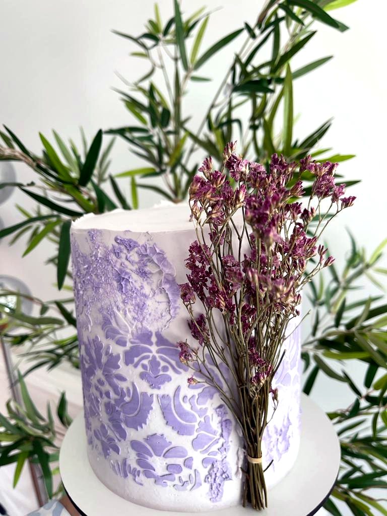 Tarta blanca y lila con textura floral, flores secas y ramas verdes alrededor.