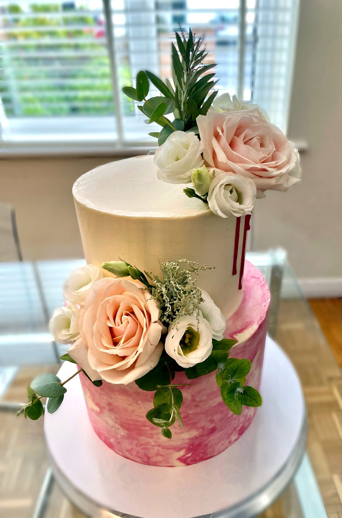 Tarta de dos niveles en blanco y rosa con rosas naturales, verdes y goteo rojo.