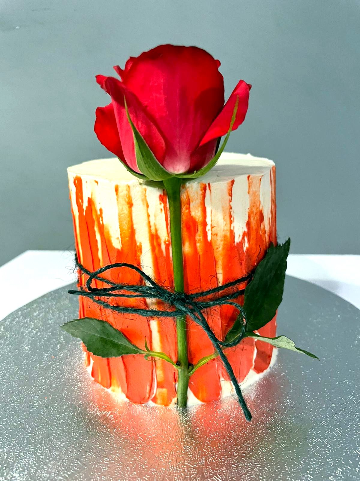 Tarta pequeña con goteo rojo, una rosa roja y lazo verde sobre base plateada.