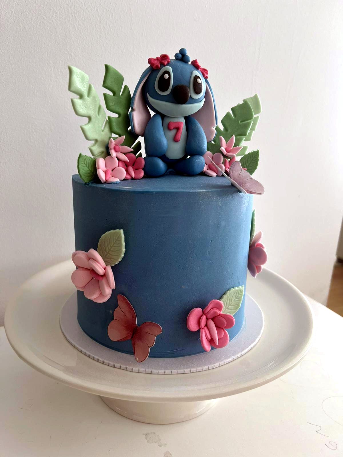 Tarta azul con figura de Stitch, flores rosas, hojas tropicales y numero siete.