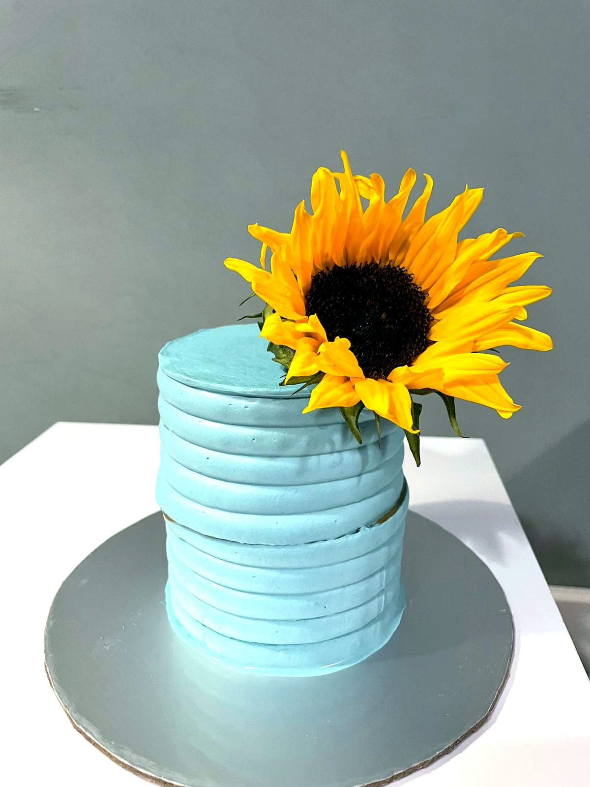 Tarta azul con textura horizontal y un girasol amarillo sobre base plateada.