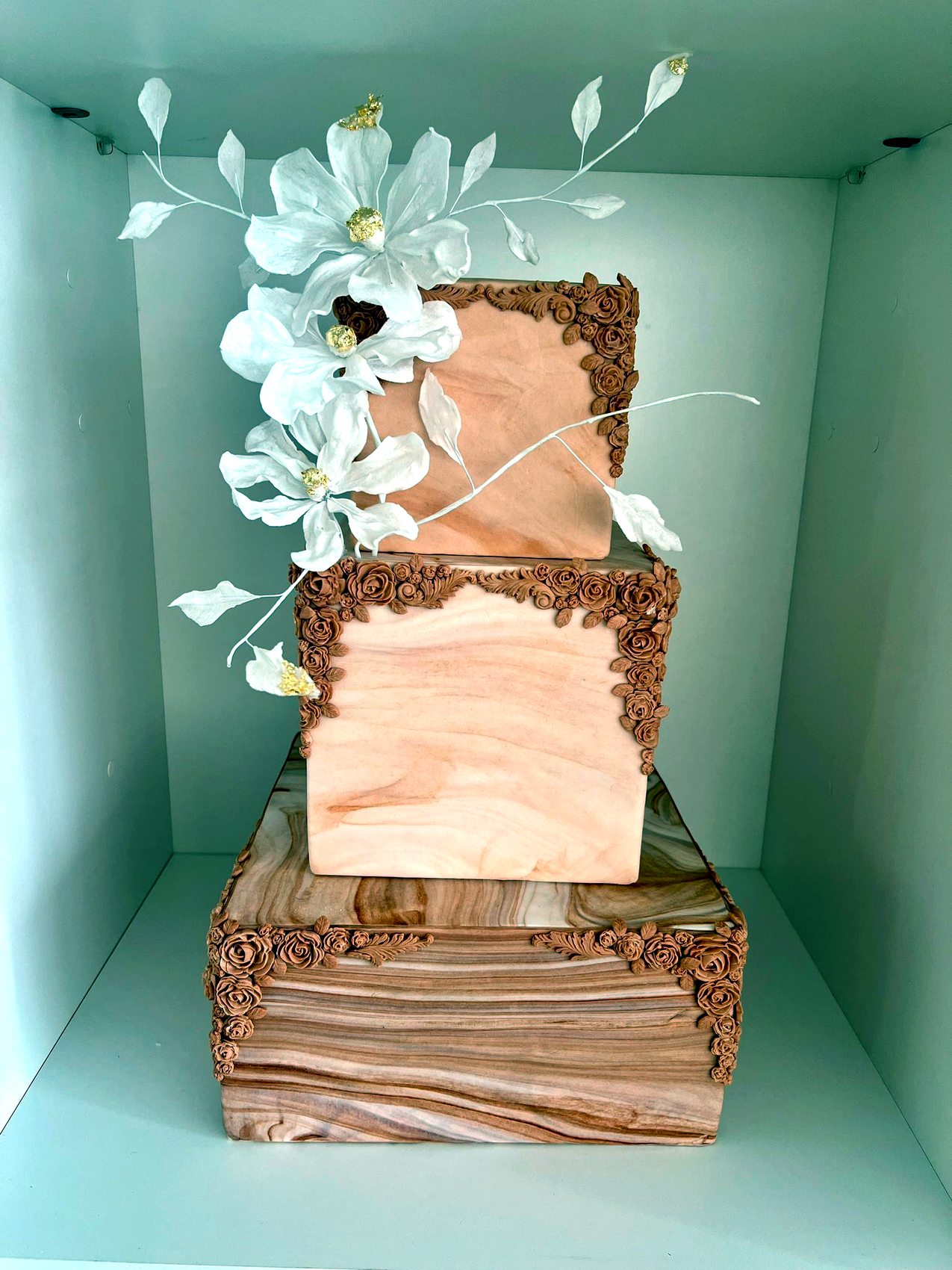 Tarta geometrica de tres niveles en tonos terracota con flores blancas y filigrana.
