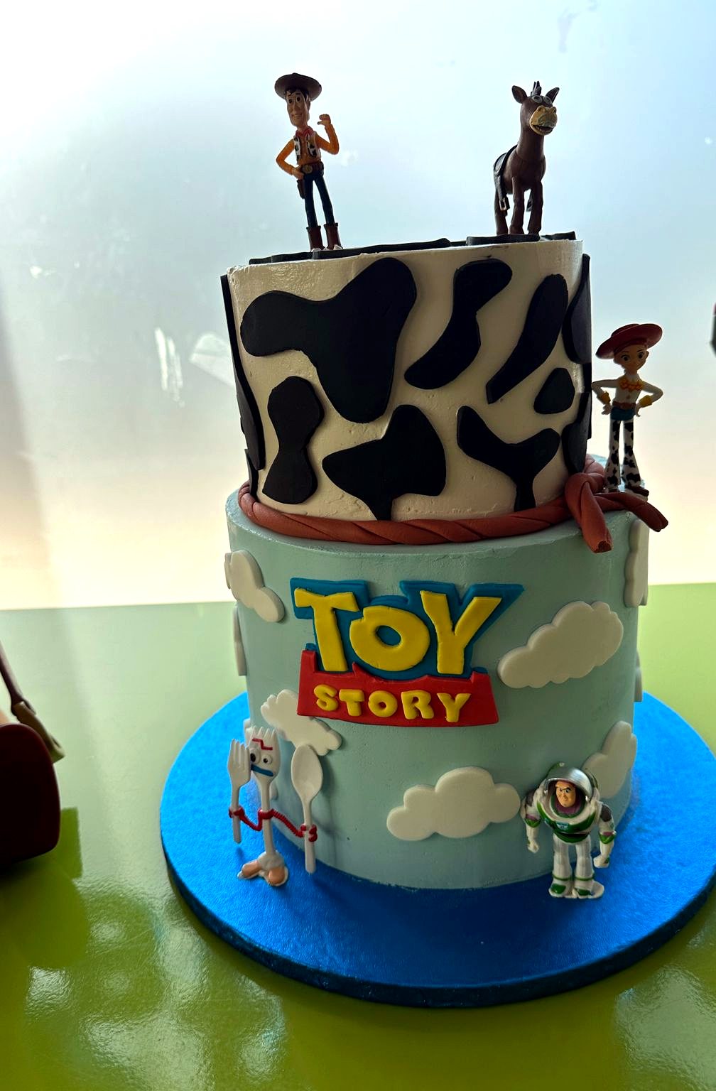 Tarta de dos niveles inspirada en Toy Story con figuras, nubes y estampado de vaca.