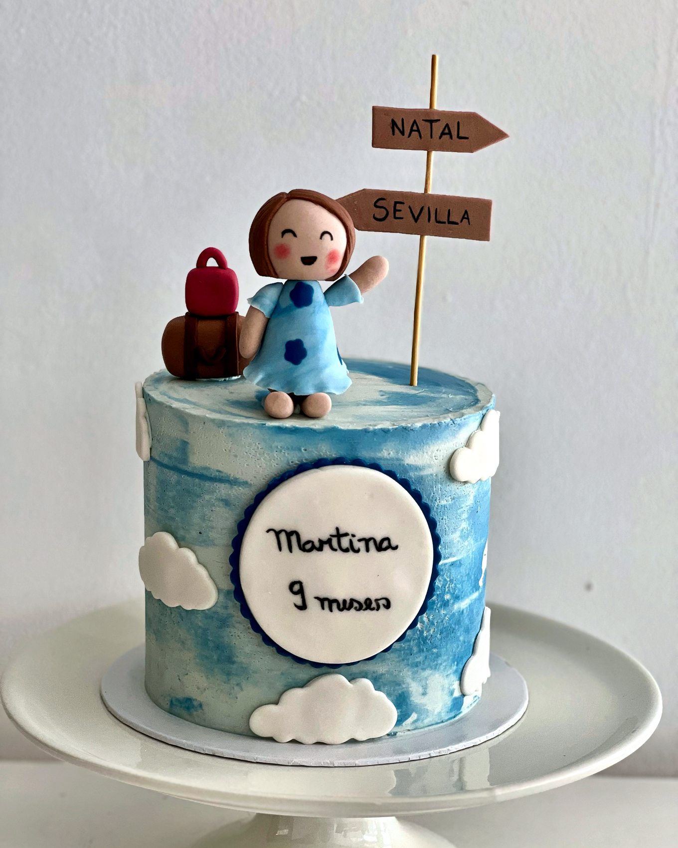 Tarta azul con figura infantil, maleta, nubes y cartel de viaje para Martina.