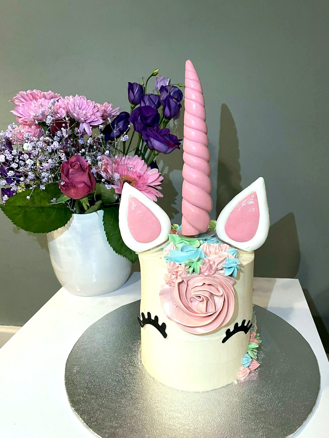 Tarta de unicornio con cuerno rosa, orejas, pestanas y flores pastel.